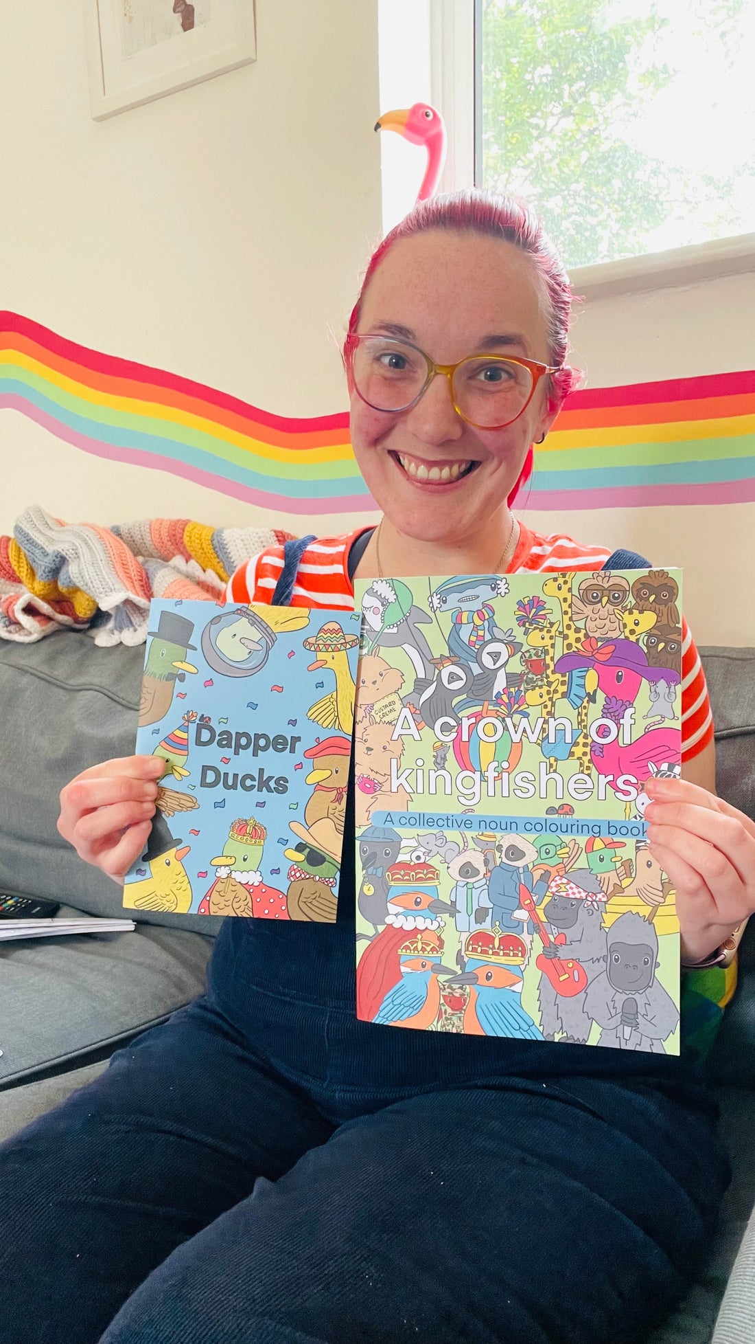 Katy is a white female with bright red hair in dark blue dungarees holding two colouring books called dapper ducks and a crown of kingfishers