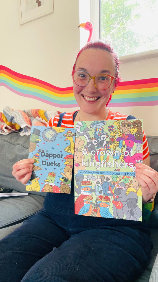 Katy is a white female with bright red hair in dark blue dungarees holding two colouring books called dapper ducks and a crown of kingfishers