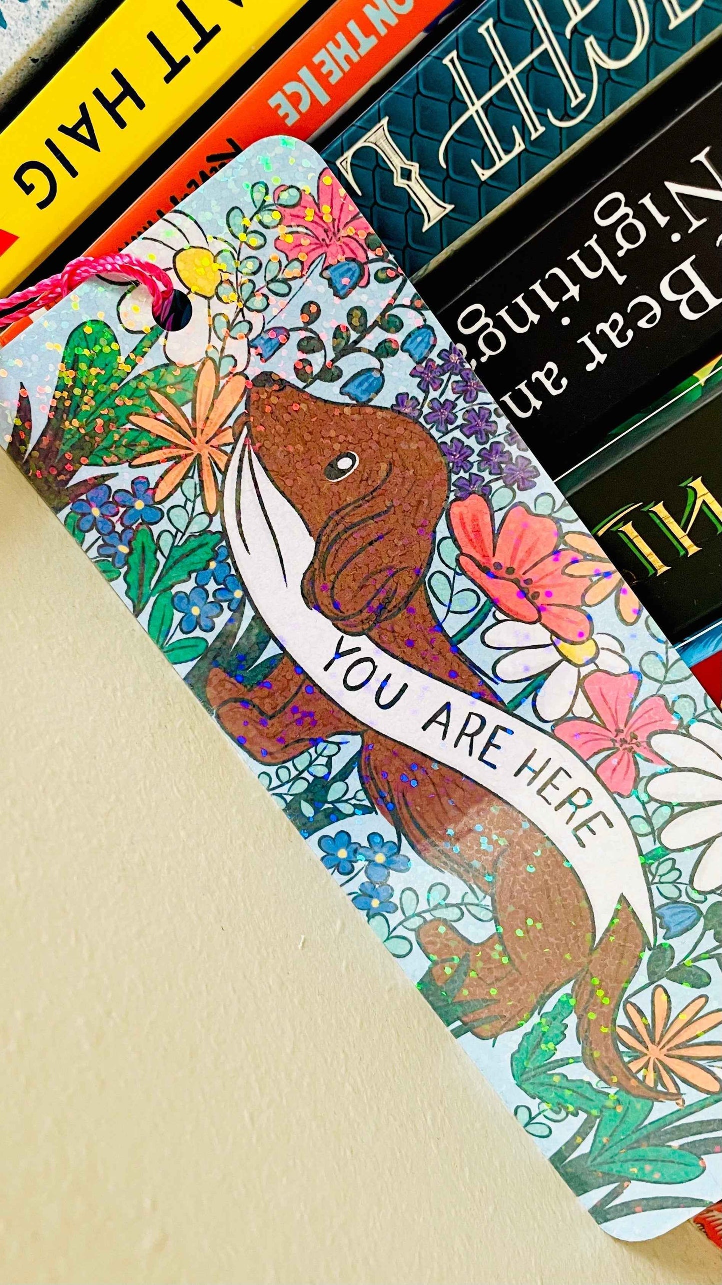 Cute handmade bookmark with a brown Dachshund surrounded by colourful wildflowers holding a sign that reads ‘you are here’. The bookmark is handmade in the UK, finished with rounded corners, rainbow holographic laminate and a coordinating tassel. Perfect for stocking fillers, gifts for dog lover, gifts for book lovers.