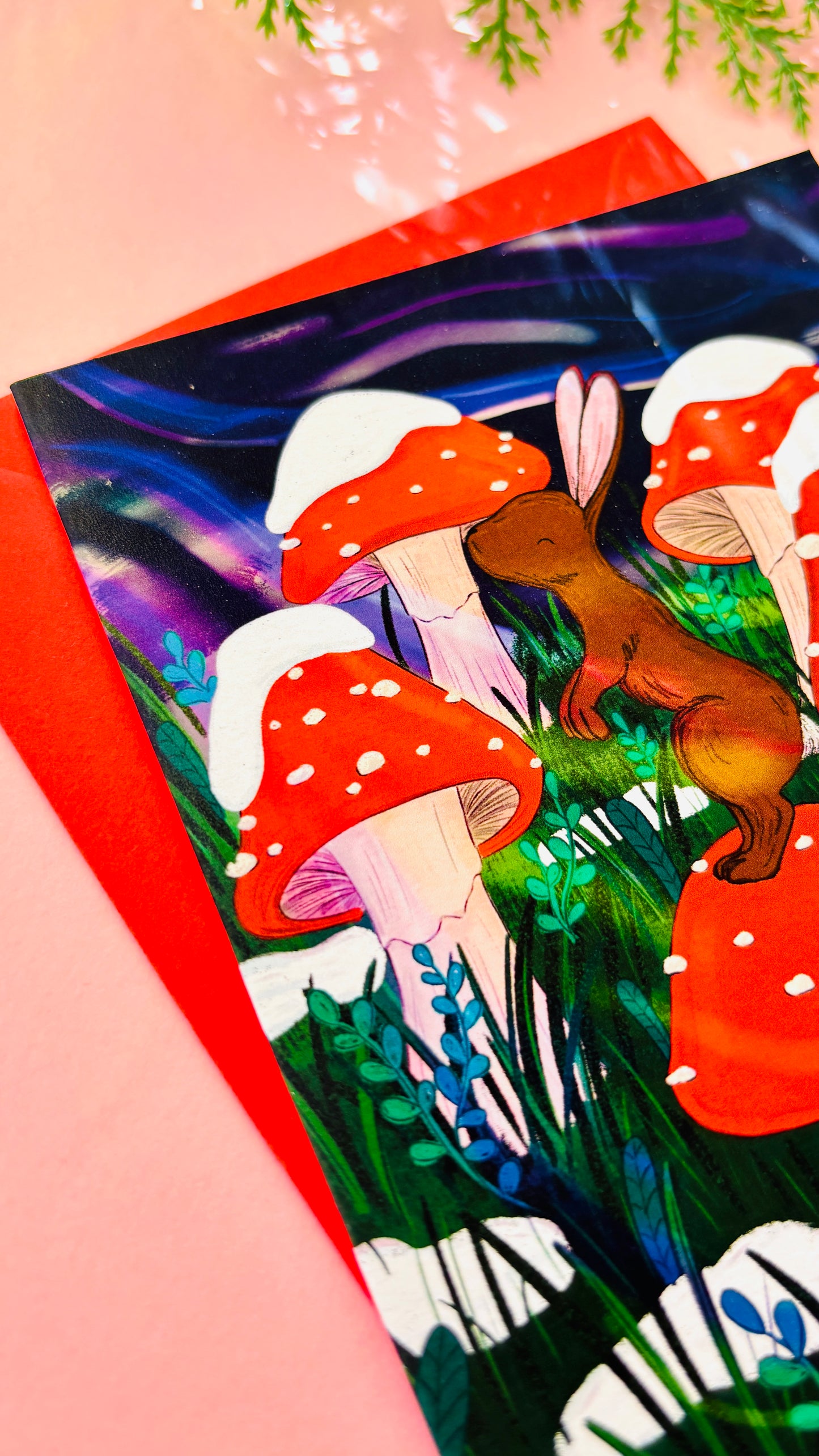 Illustrated bunny Christmas card featuring a rabbit on a toadstool surrounded by snow-dusted mushrooms and a dreamy purple sky.