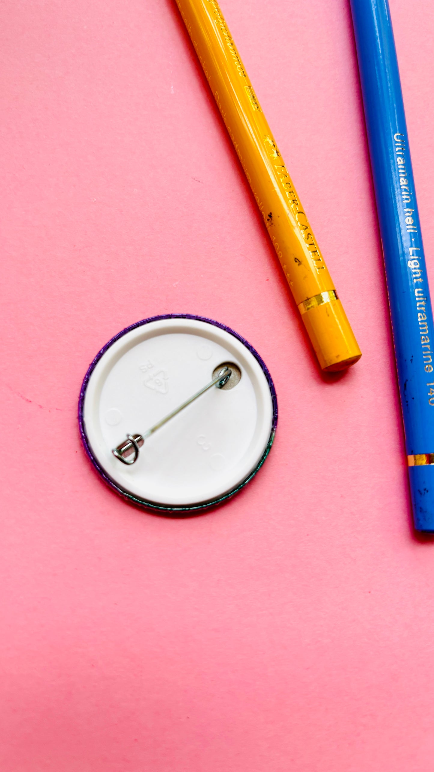 Round button badge with a pin on a pink background with two pencils.