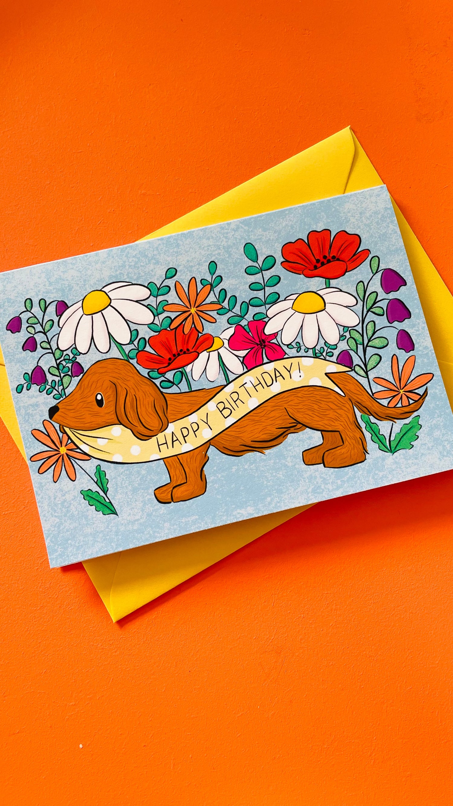 Colourful illustrated greetings card of a sausage dog holding in a yellow and white polka dot banner reading happy birthday in front a selection of wildflowers