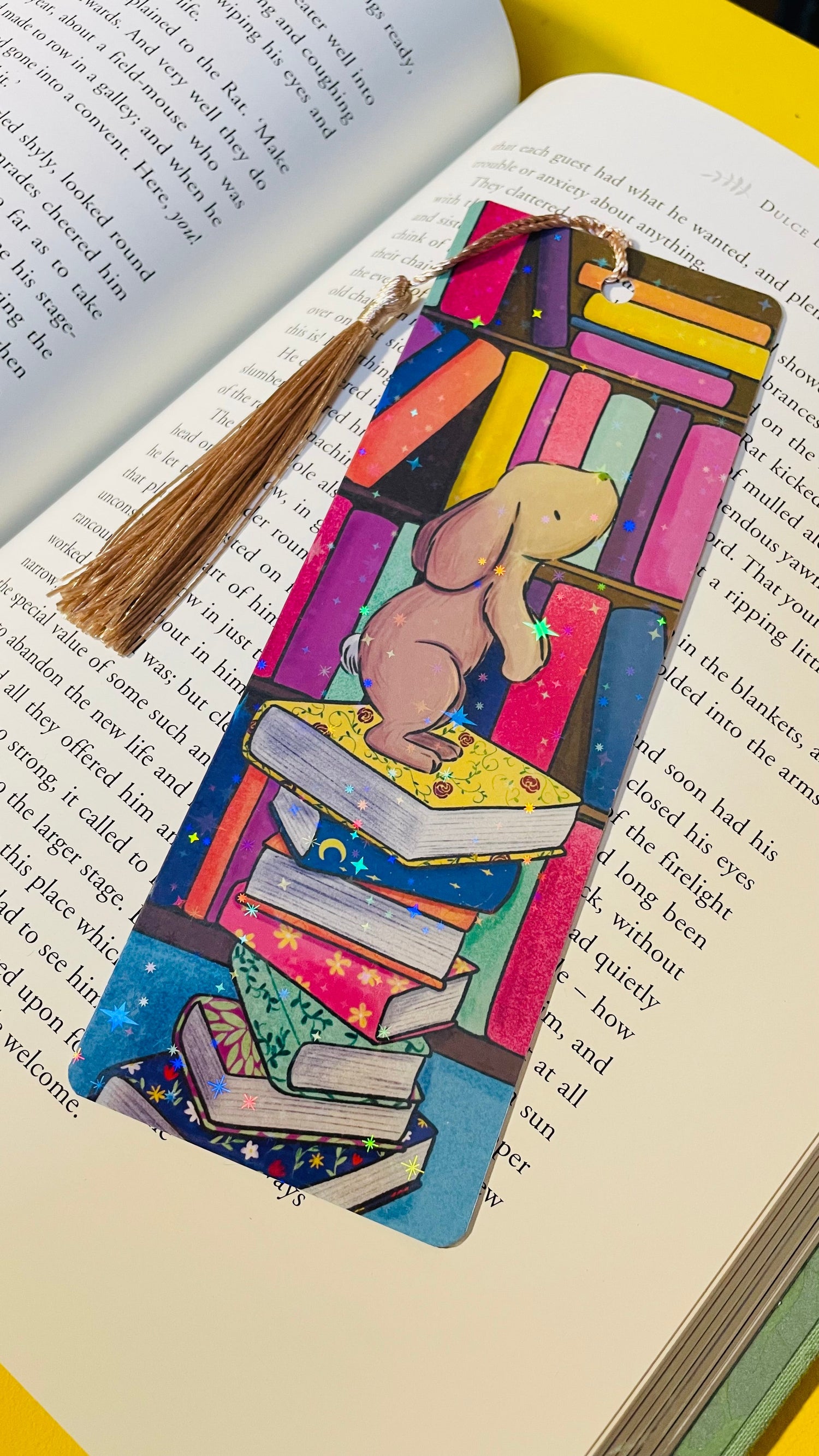 A colourful rainbow holographic bookmark of a bunny on top of a pile of books in a colourful library