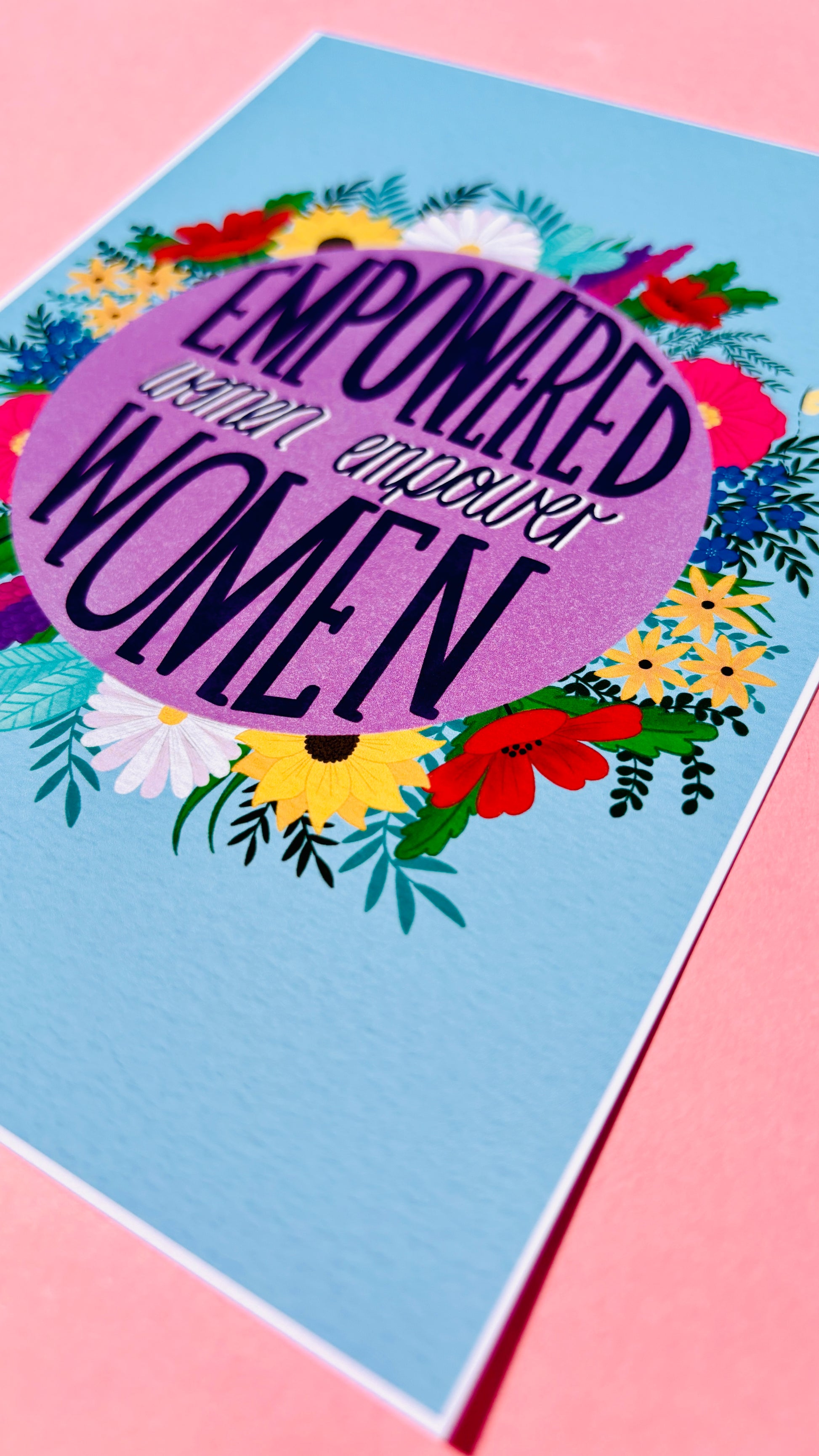 Feminist art print that reads “empowered women empower women” surround by colourful flowers and sets against a blue background. Inspirational wall art for women. 