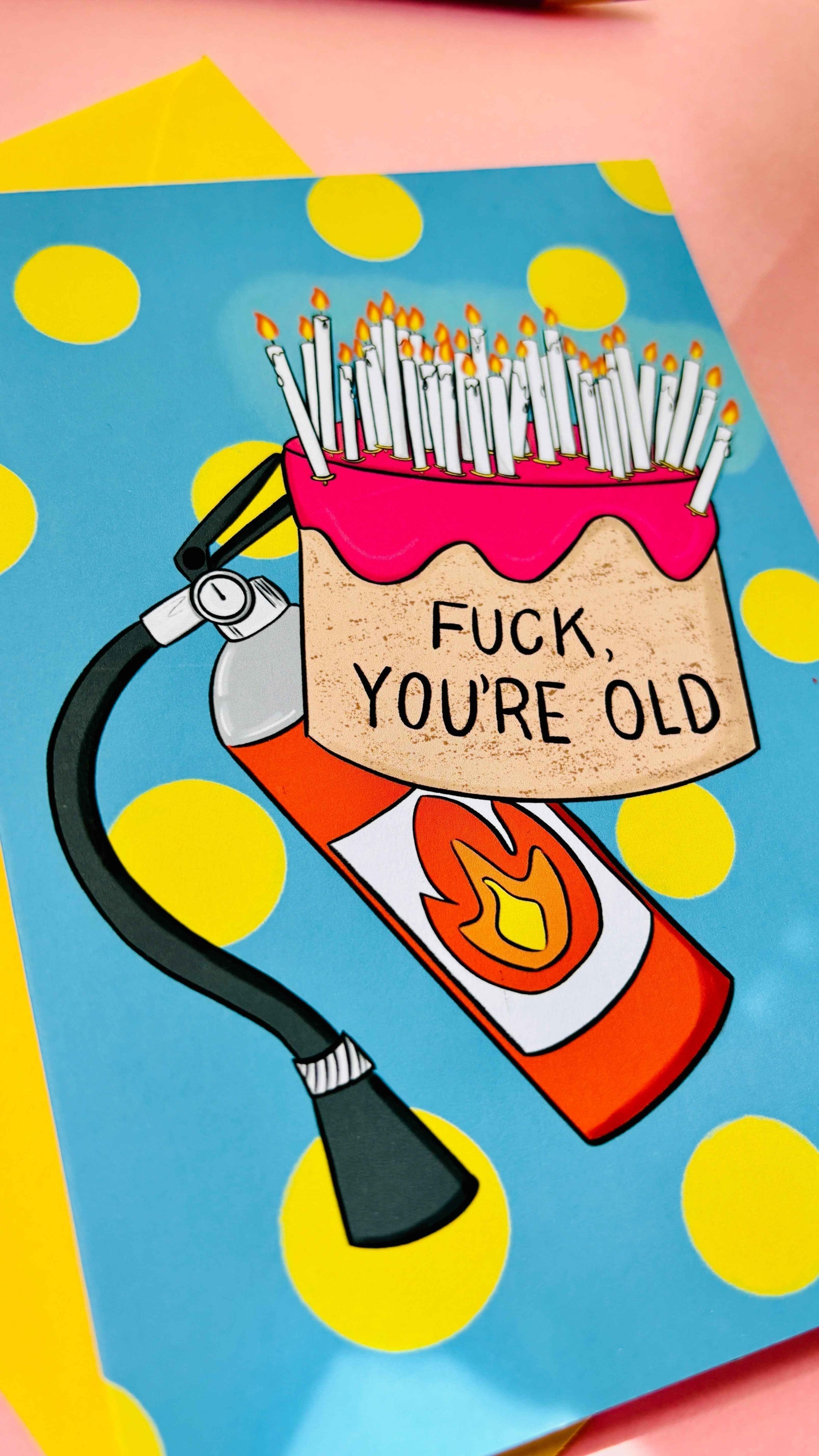 This handmade Sweary rude birthday card features a cake overflowing with candles and pre-cautionary fire extinguisher under the funny text “fuck you’re old” against a blue background with yellow polkadots 