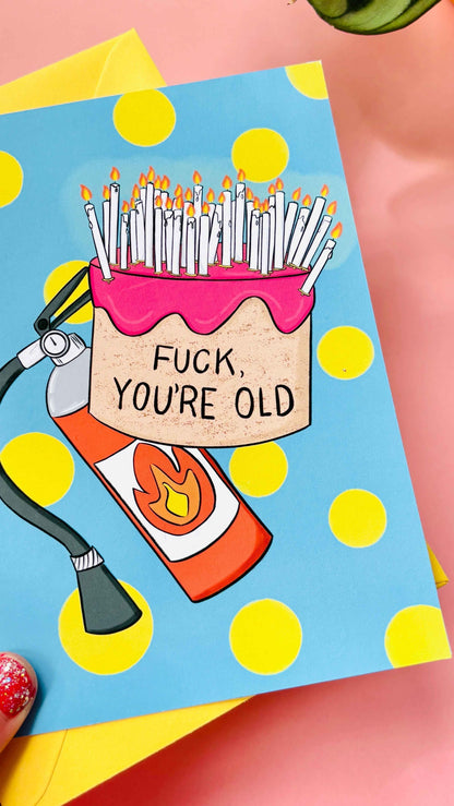 This handmade Sweary rude birthday card features a cake overflowing with candles and pre-cautionary fire extinguisher under the funny text “fuck you’re old” against a blue background with yellow polkadots 