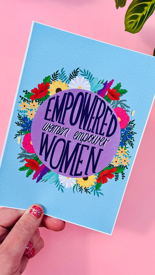 Feminist art print that reads “empowered women empower women” surround by colourful flowers and sets against a blue background. Inspirational wall art for women. 