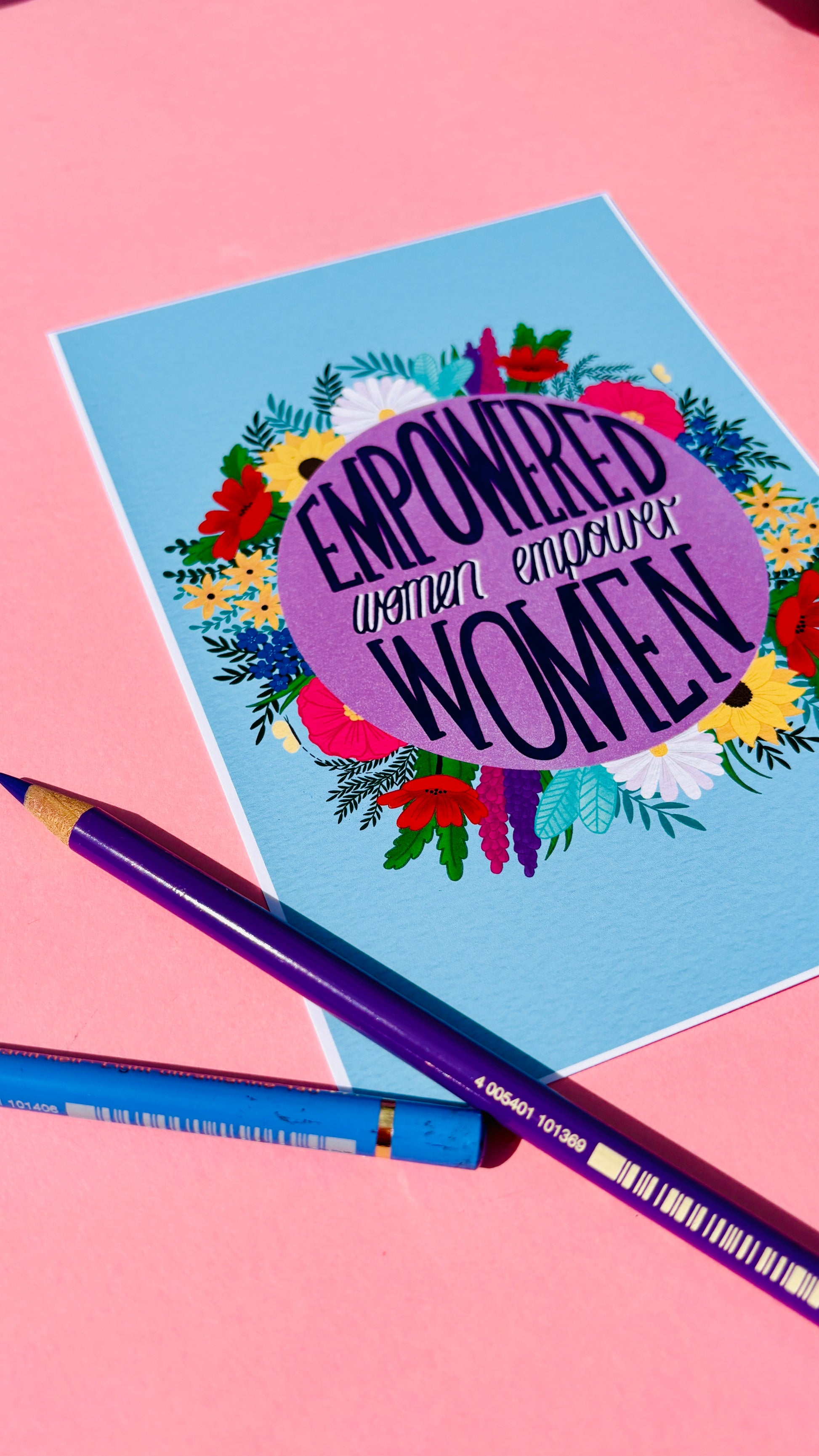 Feminist art print that reads “empowered women empower women” surround by colourful flowers and sets against a blue background. Inspirational wall art for women. 