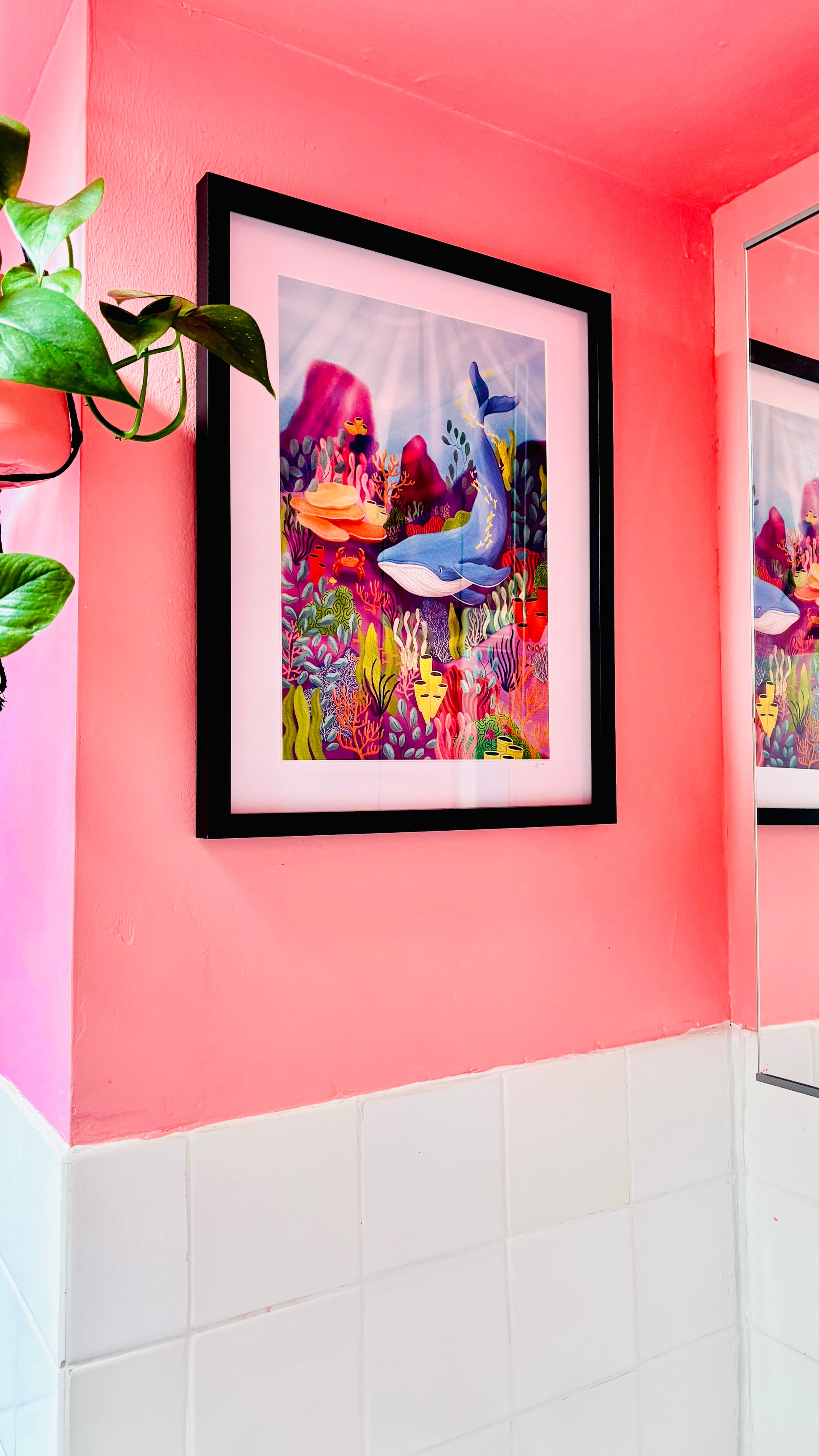 A colourful art print of a coral reef and blue whale scene framed on a pink bathroom wall 