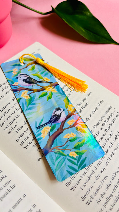Handmade bookmark with illustration of two cute long tailed tits sitting on delicate branches surrounded by yellow flowers. Finished with rainbow holographic laminate, rounded corners and a tassel. Ideal gift for bookish gifts lovers. 