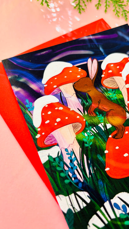 Illustrated bunny Christmas card featuring a rabbit on a toadstool surrounded by snow-dusted mushrooms and a dreamy purple sky.