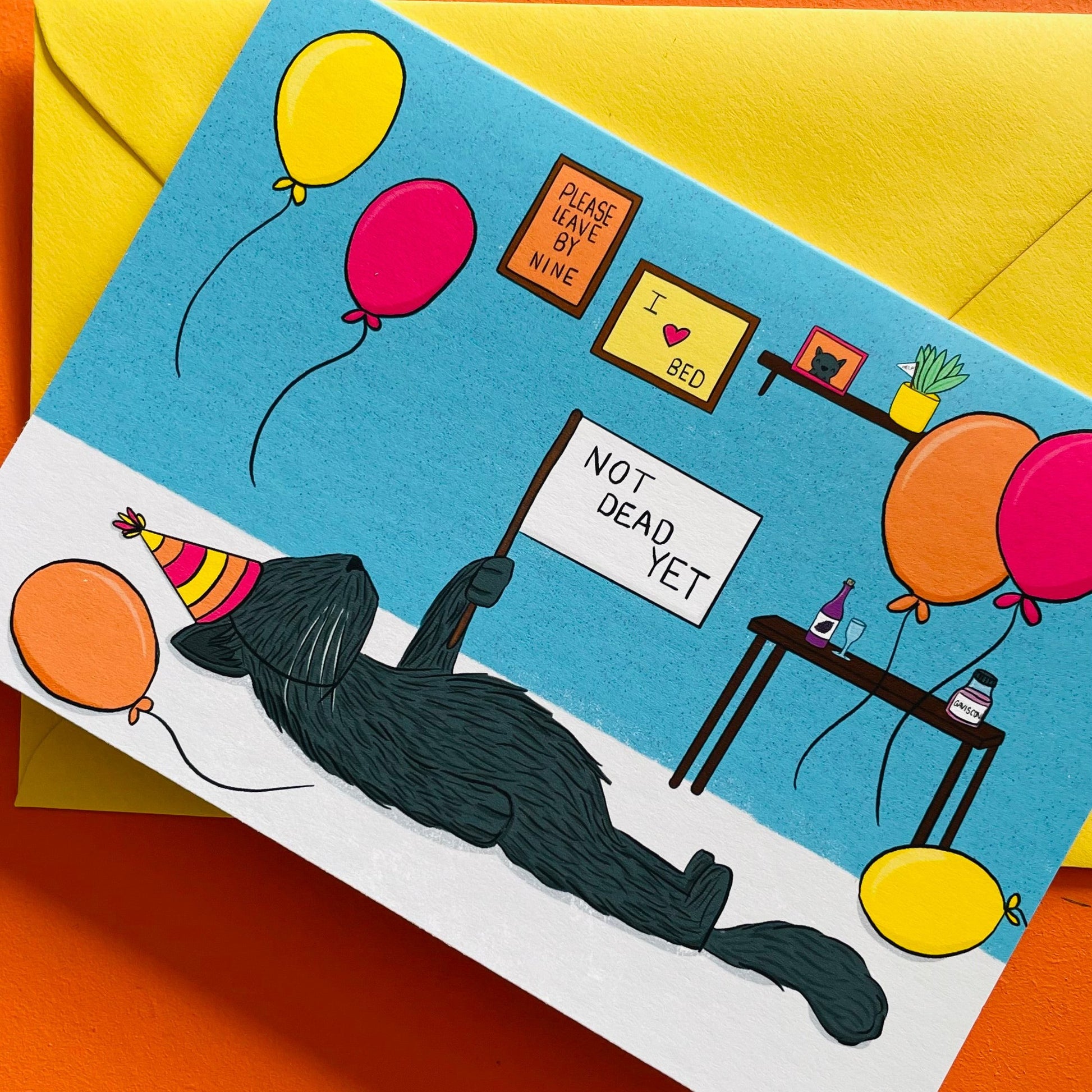 Illustrated birthday card featuring a grey kitty lying on the floor in a party hat holding a flag that reads “not dead yet”