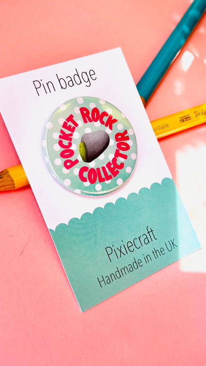 A round pin badge reading 'POCKET ROCK COLLECTOR' with a small dark stone in the center, mounted on a branded Pixiecraft display card on a pink background with colored pencils. Handmade accessory for nature lovers and geology gifts