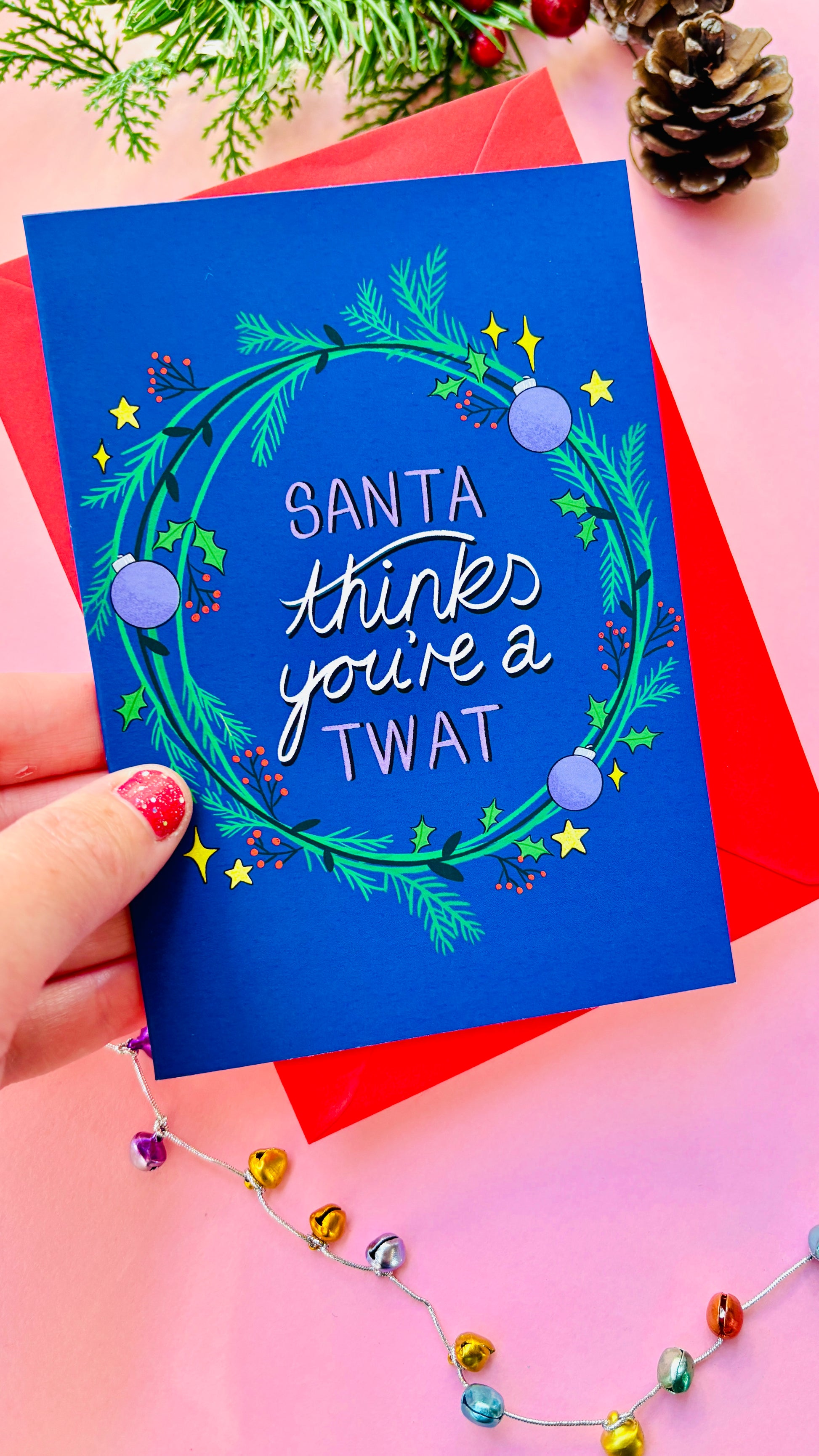 Sweary Christmas card reading “Santa thinks you’re a twat” surrounded by green and purple wreath on a blue background.