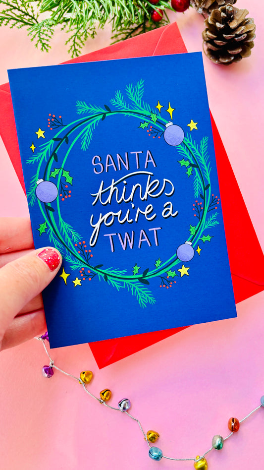 Sweary Christmas card reading “Santa thinks you’re a twat” surrounded by green and purple wreath on a blue background.