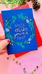 Sweary Christmas card reading “Santa thinks you’re a twat” surrounded by green and purple wreath on a blue background.