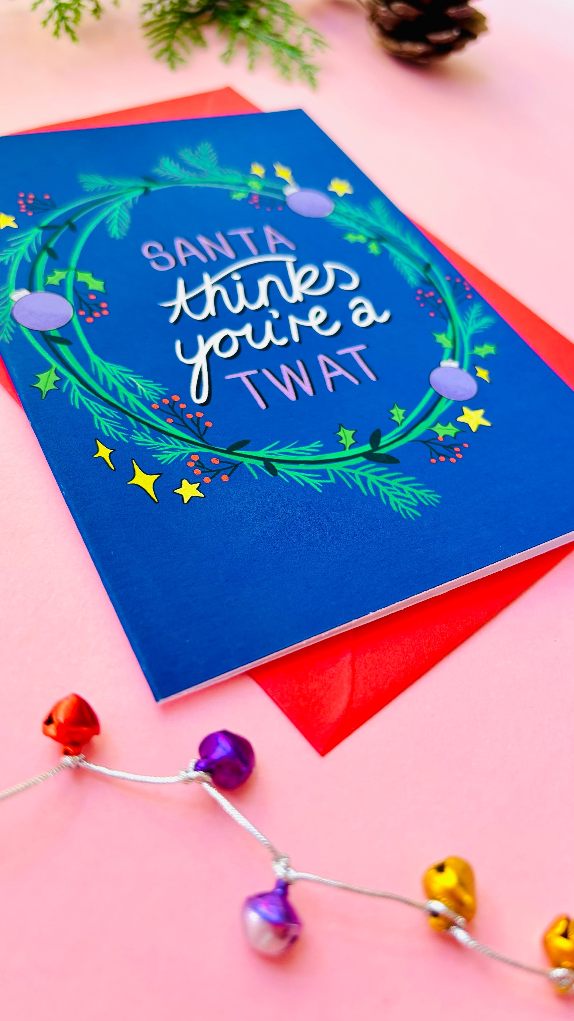 Funny adult Christmas card featuring the phrase “Santa thinks you’re a twat” framed by a green and purple wreath on a deep blue background.