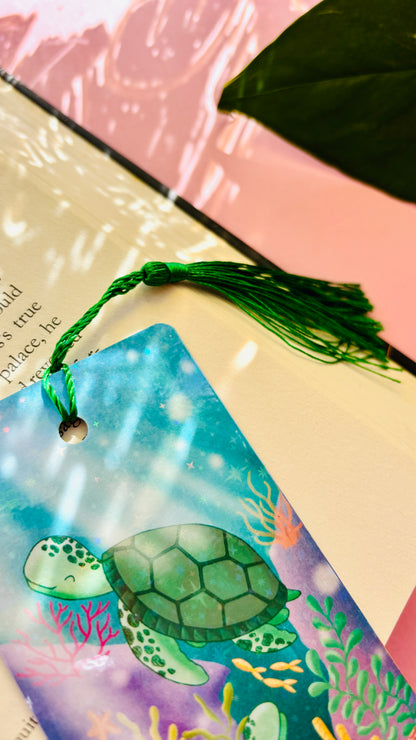 Double-sided turtle gift bookmark showing two hand-drawn sea turtles swimming in a coral reef, finished with rainbow holographic laminate and green tassel for marking pages.