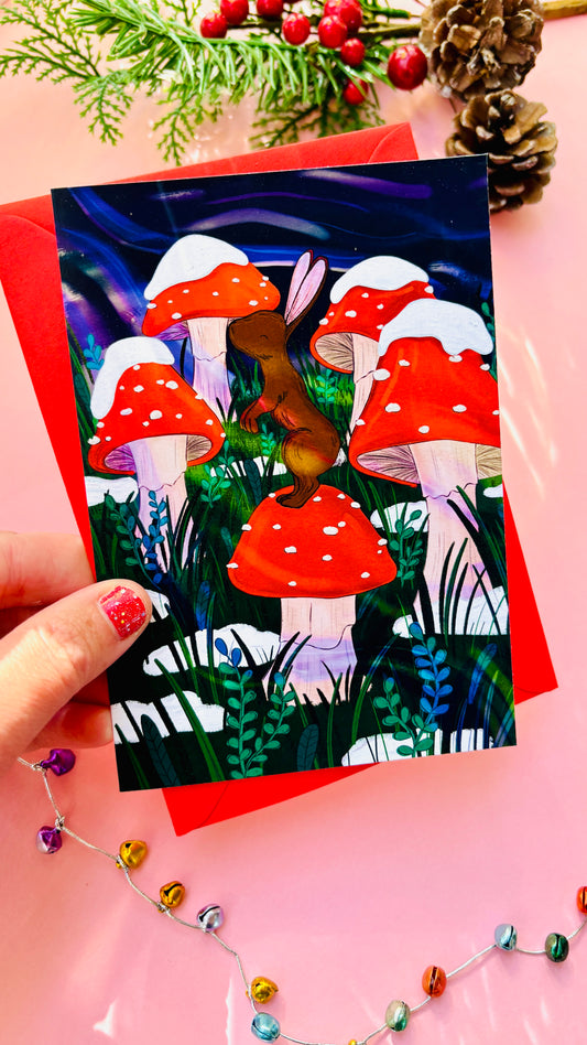 Handmade woodland Christmas card with a brown bunny on a red toadstool under a magical winter sky, perfect for nature lovers.