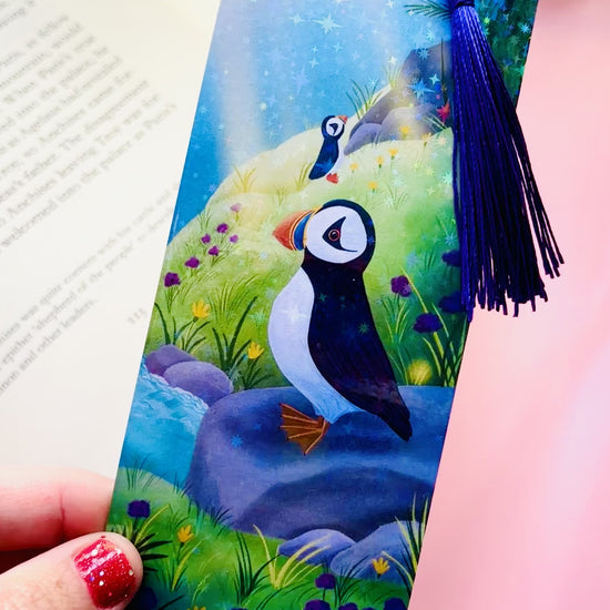 Handmade puffin bookmark with rainbow holographic finish, featuring a cute puffin on a colorful coastal seaside scene, FSC-certified paper, and coordinating tassel, perfect for book lovers and bird enthusiasts.
