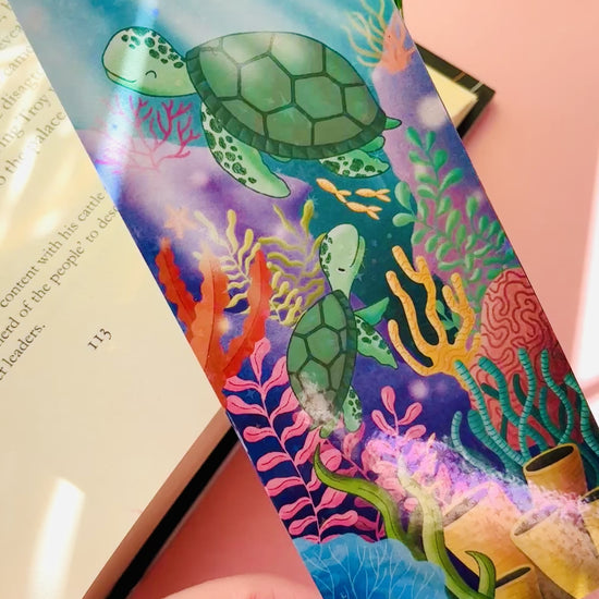 Ocean-inspired bookmark featuring two cute sea turtles swimming through a colorful coral reef, double-sided with rainbow holographic laminate and green tassel, perfect for readers and marine life fans.