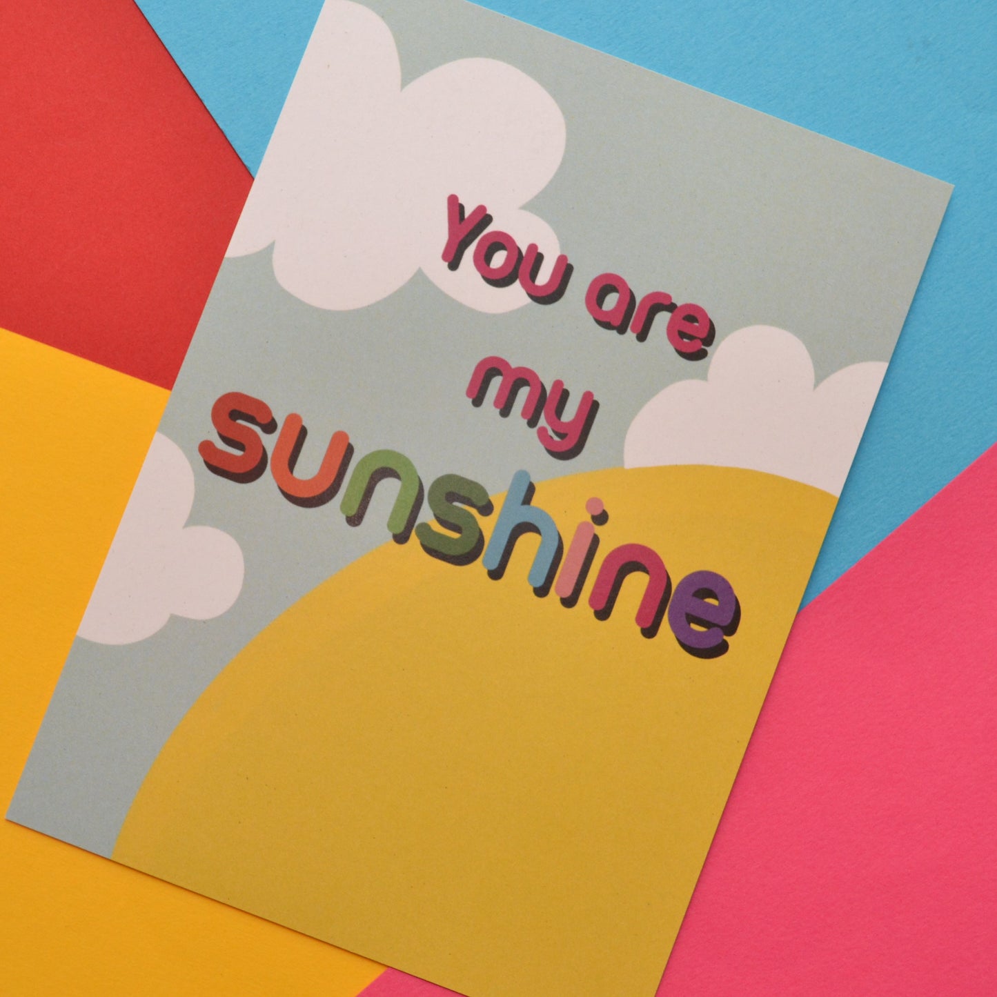 You are my sunshine postcard in red and rainbow colours. The background is a bright yellow sun, blue background and white clouds.