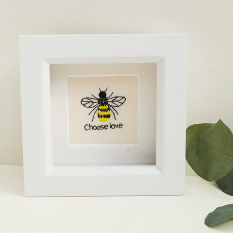 A miniature hand embroidered bee above the text choose love. Framed in a white box frame