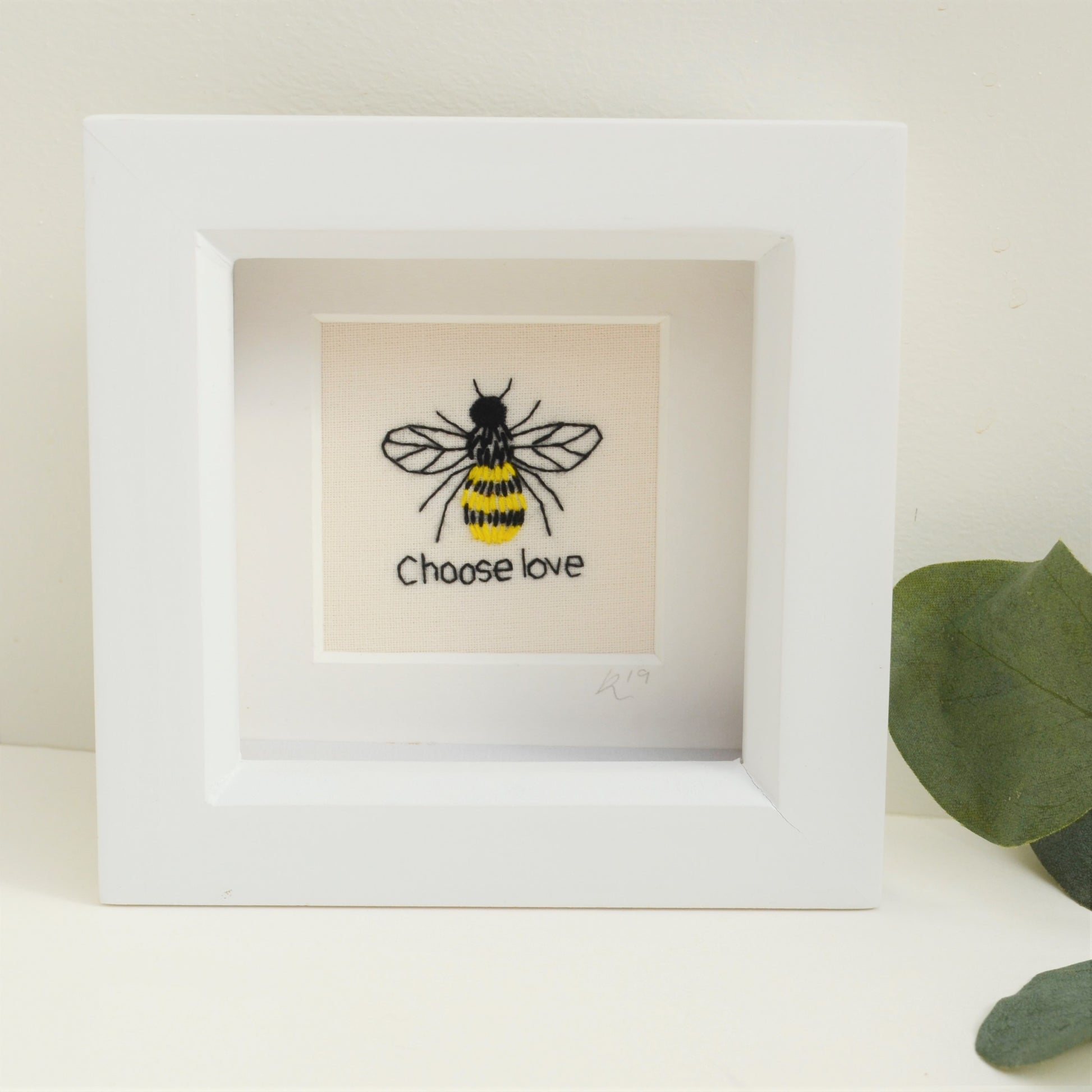 A small embroidered bee and the text reads choose love. It is framed in a deep set white box frame