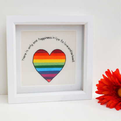 A white box frame next to a colourful flower. The embroidery is a rainbow striped heart and the text "There is only one happiness in life: to love and be loved"