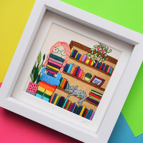 An original piece of mixed media hand embroidery. This pieces shows a fair skinned girl with long bright red hair and blue glasses holding a pile of colourful books. She is wearing a cosy textured jumper and rainbow leggings. Behind her is a tall bookcase full of colourful books. On the bookcase is a trailing green plant in a pink pot. Asleep on the bookcase shelf is a grey cat. On the other side of the girl is a tall plant in a pink and grey plant pot. 