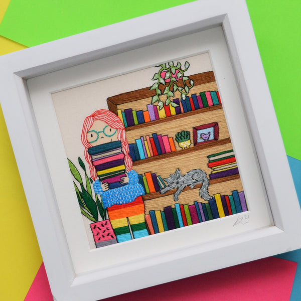 An original piece of mixed media hand embroidery. This pieces shows a fair skinned girl with long bright red hair and blue glasses holding a pile of colourful books. She is wearing a cosy textured jumper and rainbow leggings. Behind her is a tall bookcase full of colourful books. On the bookcase is a trailing green plant in a pink pot. Asleep on the bookcase shelf is a grey cat. On the other side of the girl is a tall plant in a pink and grey plant pot. 