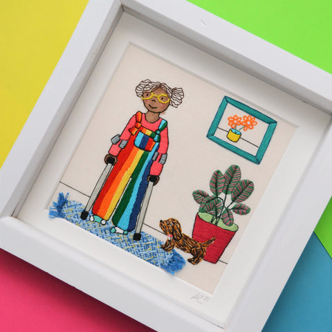 This pieces shoes a black girl wearing bright rainbow dungarees. She has brown curly hair, bright yellow glasses and odd socks. She is leaning on a pair of crutches and is stood alongside a large calathea plant and brown speckled sausage dog. On the wall is a green frame with a picture of a yellow pot with orange flowers in and on the floor is a textured weaved blue rug with tiny tassels.