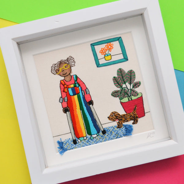 This pieces shoes a black girl wearing bright rainbow dungarees. She has brown curly hair, bright yellow glasses and odd socks. She is leaning on a pair of crutches and is stood alongside a large calathea plant and brown speckled sausage dog. On the wall is a green frame with a picture of a yellow pot with orange flowers in and on the floor is a textured weaved blue rug with tiny tassels.