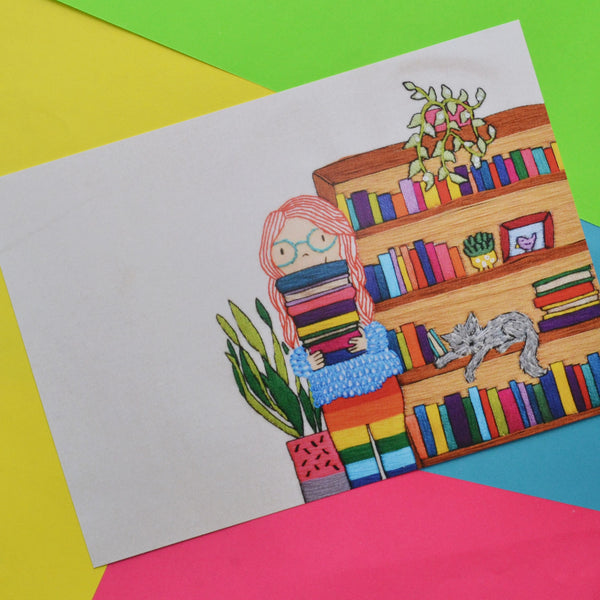 An A5 print of the original mixed media hand embroidery. This pieces shows a fair skinned girl with long bright red hair and blue glasses holding a pile of colourful books. She is wearing a cosy textured jumper and rainbow leggings. Behind her is a tall bookcase full of colourful books. On the bookcase is a trailing green plant in a pink pot. Asleep on the bookcase shelf is a grey cat. On the other side of the girl is a tall plant in a pink and grey plant pot. 