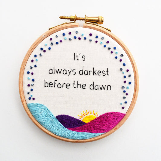 hand embroidery hoop art features the uplifting quote "It's always darkest before the dawn" above purple and blue rolling hills and and a rising sun underneath a french knot sky.