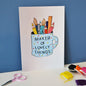 An a5 print of the maker of lovely things mug hand embroidery. The print shows the blue polka dot mug which reads maker of lovely things full of pens, pencils, paintbrushes and a ruler. The print is stood against a dark blue background next to some small scissors and bobbins of thread