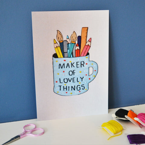 An a5 print of the maker of lovely things mug hand embroidery. The print shows the blue polka dot mug which reads maker of lovely things full of pens, pencils, paintbrushes and a ruler. The print is stood against a dark blue background next to some small scissors and bobbins of thread