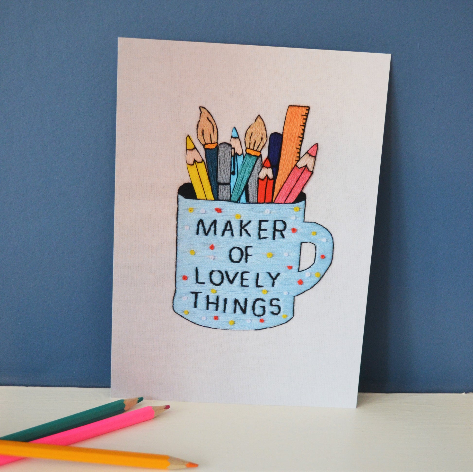 An a5 print of a hand embroidery. It shows a polka dot blue mug which has the words maker of lovely things in black text. Inside the mug are a selection of colourful pens, pencils, paintbrushes and a ruler. The print is propped up against a dark blue background alongside a selection of pencils. 