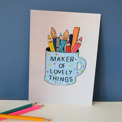 An a5 print of a hand embroidery. It shows a polka dot blue mug which has the words maker of lovely things in black text. Inside the mug are a selection of colourful pens, pencils, paintbrushes and a ruler. The print is propped up against a dark blue background alongside a selection of pencils. 