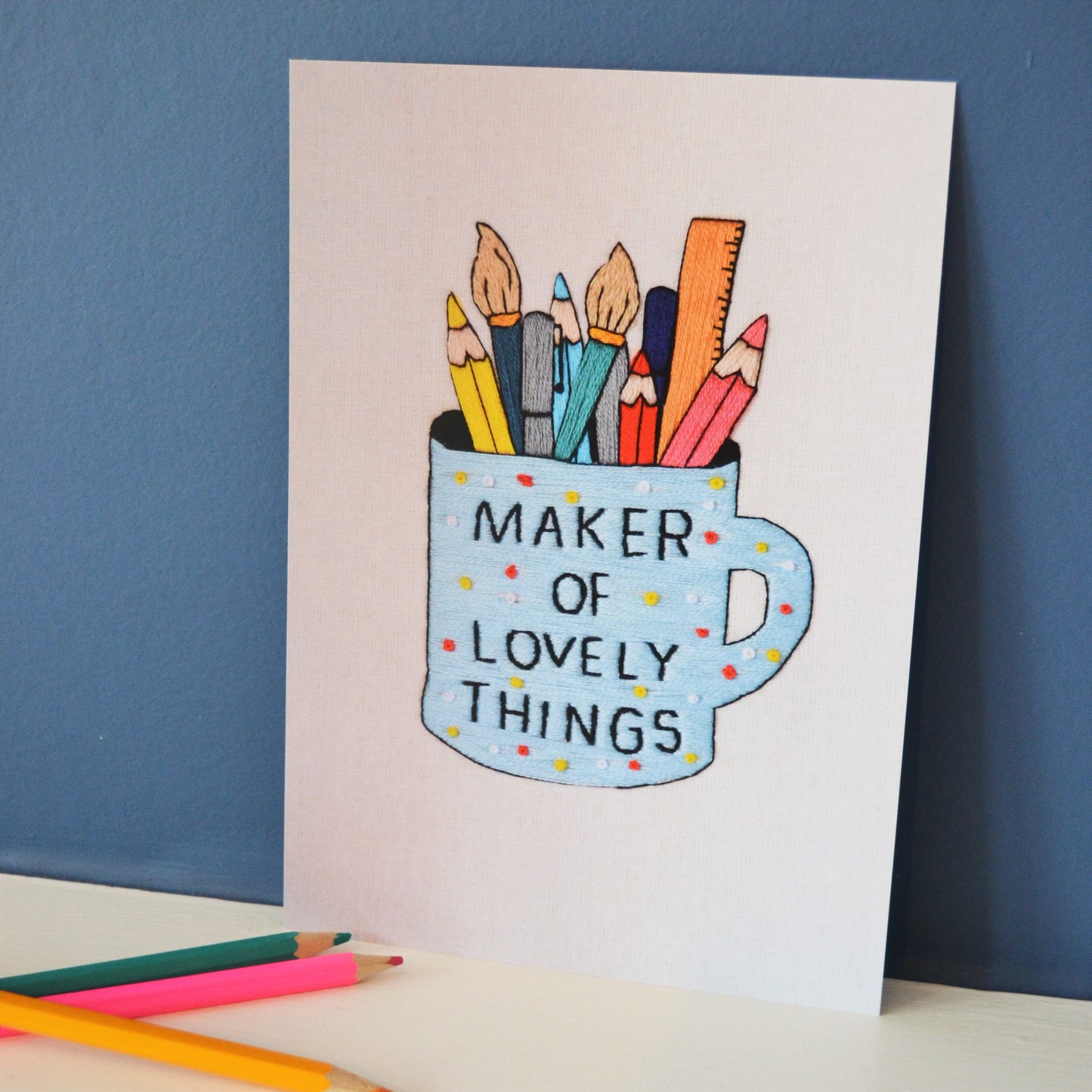 An A5 print of a piece of hand embroidery. The embroidery is of a mug filled with pens pencils paintbrushes and a ruler. The mug reads maker of lovely things. The print is propped up against a blue wall next to a few coloured pencils.