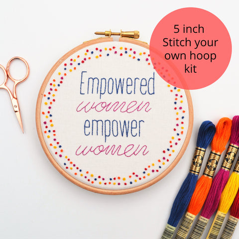 Hand Embroidery hoop reading empowered women empower women in navy blue and violet. It is surrounded by colourful french knots. Alongside the hoop are some rose gold embroidery scissors and a few colour skeins of thread